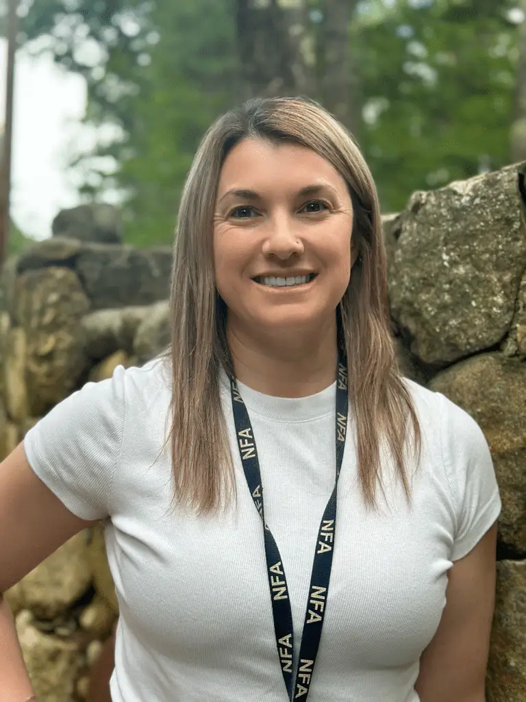 Smiling woman wearing an NFA lanyard and white T-shirt by a stone wall